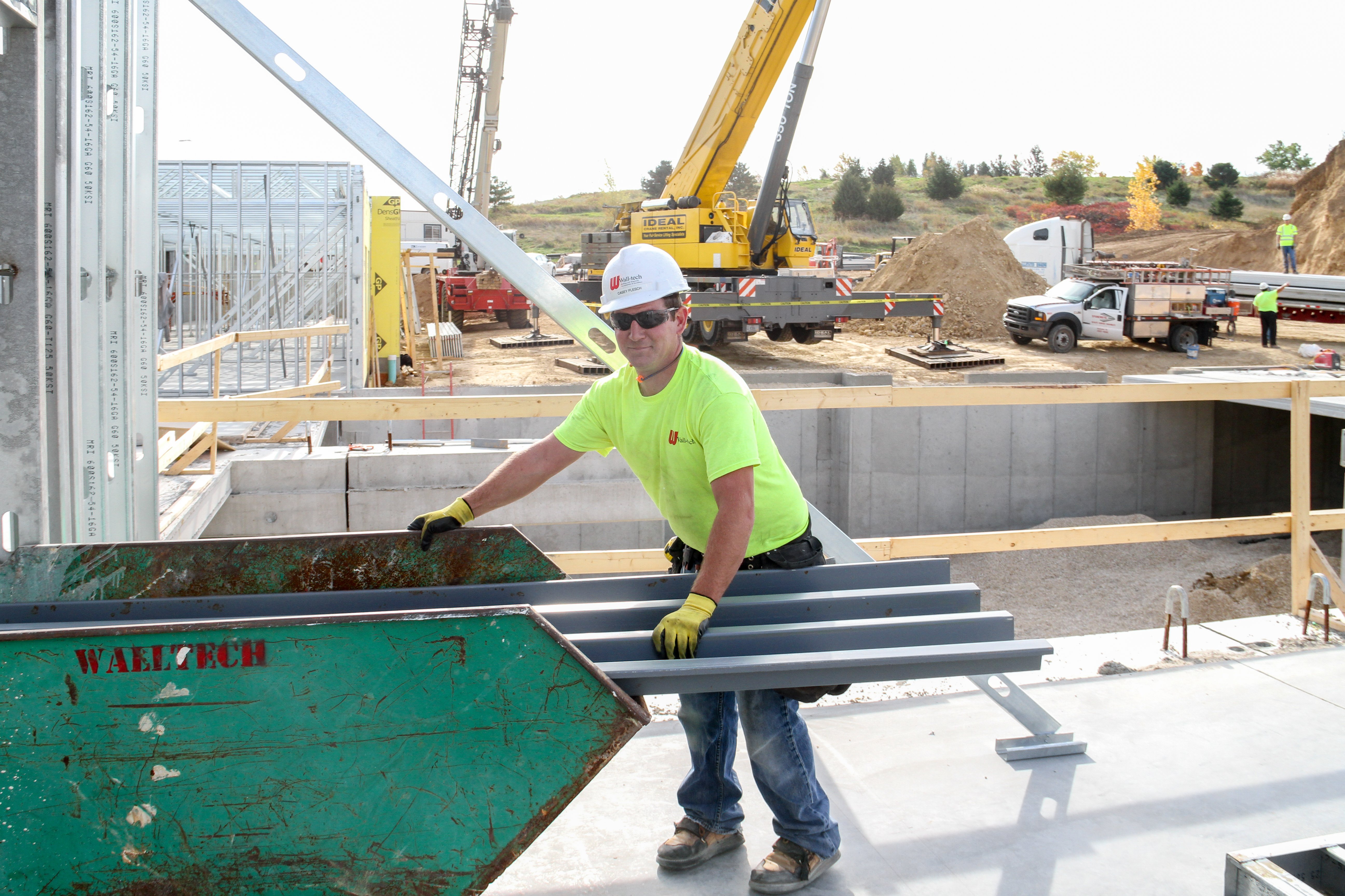 Worker carrying a steel framing member at a cold-formed steel panel job site with a crane in the background.